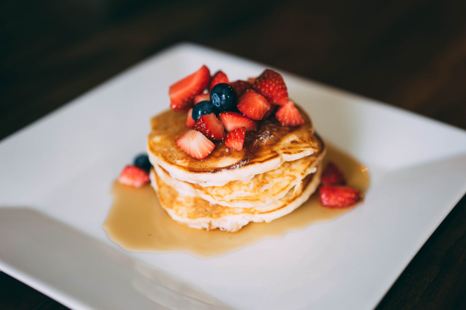 Stack of pancakes topped with strawberries and blueberries in a pool of syrup on a white plate